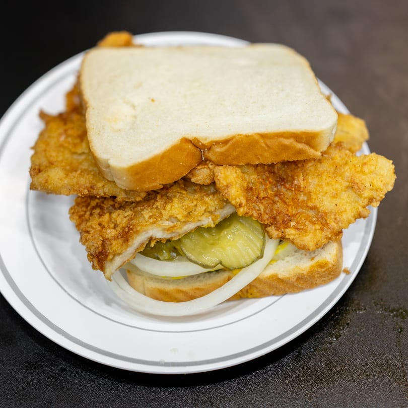 Fried fish sandwich with pickles, onions, and mustard on white bread, served on a white plate.
