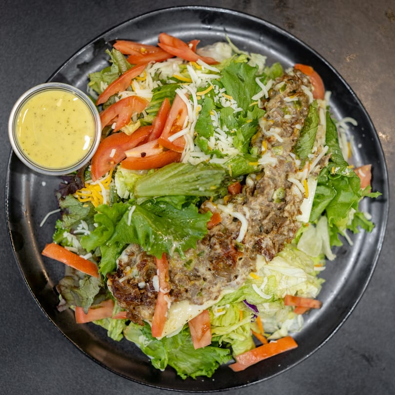 A large salad with mixed greens, shredded cheese, sliced tomatoes, and a hamburger patty, with dressing on the side.
